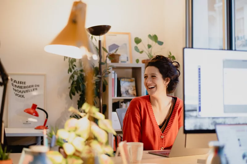 Une personne de l’équipe rit à son bureau, affiche « Faire émerger de nouvelles voix » au mur.